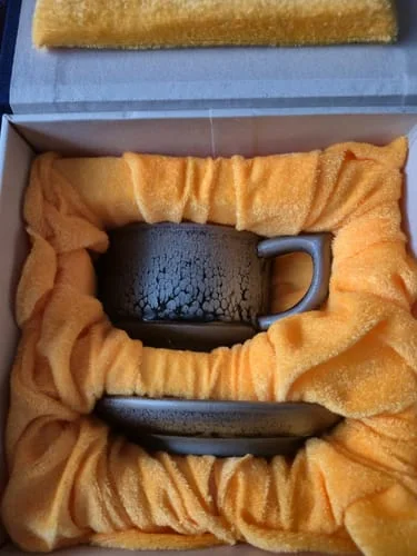 Customer's Raindrops Coffee Cup and saucer with a speckled glaze, nestled in orange fabric inside a gift box.